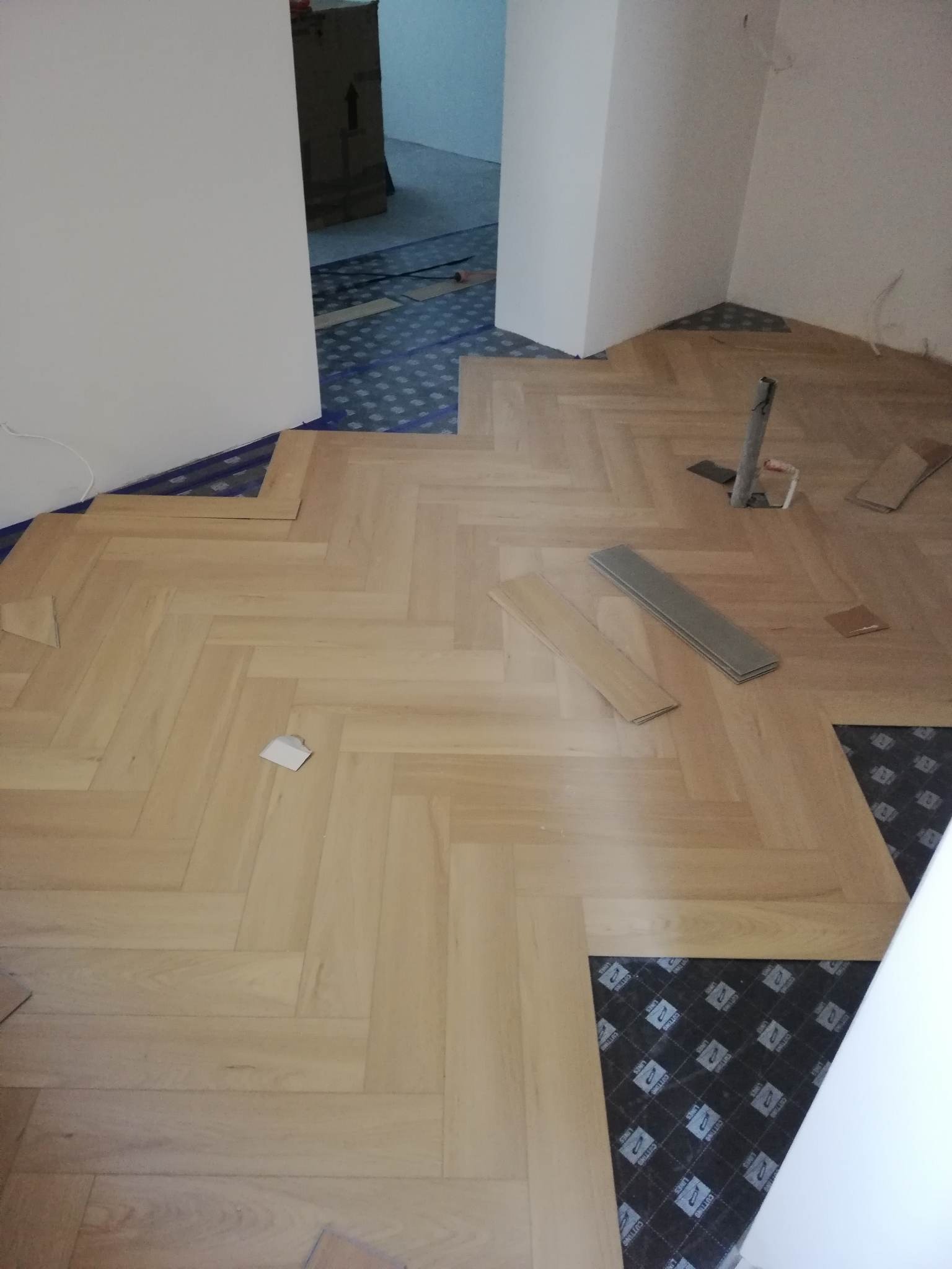 Układanie jasnych paneli podłogowych w jodełkę, widoczne niedokończone fragmenty oraz podkład izolacyjny, fragment ściany w tle.