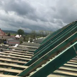 Surowa konstrukcja drewniana dachu w trakcie montażu, widok z góry na elementy nośne i łaty, w tle zabudowa wiejska i pochmurne niebo.