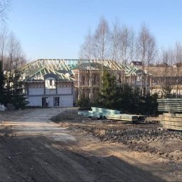 Usługi Ciesielskie Albin Gojtowski - Konstrukcja drewniana dachu w trakcie budowy domu jednorodzinnego, widok z ziemi na budynek w stanie surowym otwartym, z przygotowanym materiałem budowlanym na pierwszym planie.