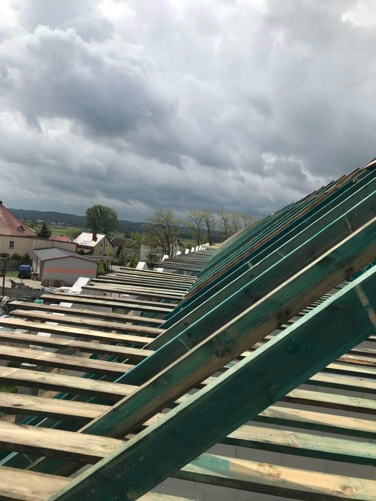 Surowa konstrukcja drewniana dachu w trakcie montażu, widok z góry na elementy nośne i łaty, w tle zabudowa wiejska i pochmurne niebo.