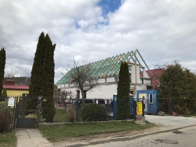 Budowa domu jednorodzinnego w trakcie realizacji, widoczna konstrukcja dachu z zielonymi elementami, ściany z białego bloczka, kontener budowlany na posesji.