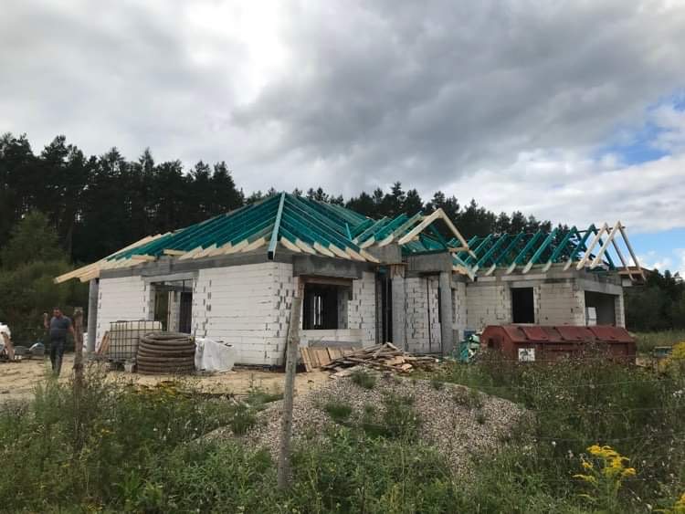 Budowa domu jednorodzinnego w stanie surowym otwartym: wzniesione ściany z bloczków, częściowo wykonana konstrukcja dachu z zieloną membraną, widoczne elementy drewniane, w tle las.