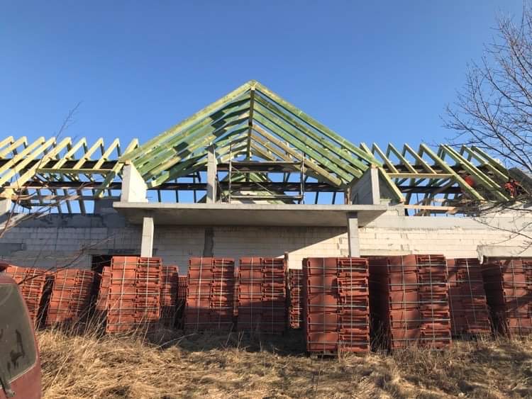 Konstrukcja drewniana dachu w trakcie budowy, widoczne przygotowane dachówki na paletach, budynek w stanie surowym otwartym.