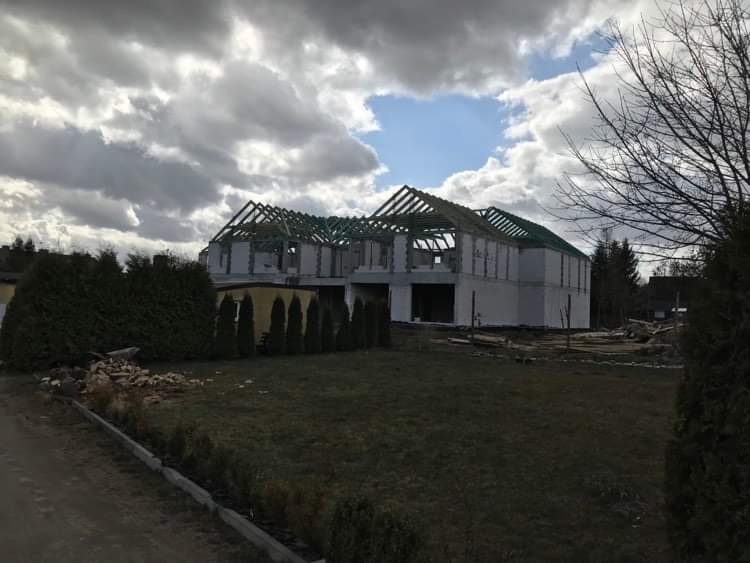 Surowy stan domu w trakcie budowy z widoczną konstrukcją dachu, białe ściany z betonu komórkowego i zielone elementy zabezpieczające, na tle pochmurnego nieba.