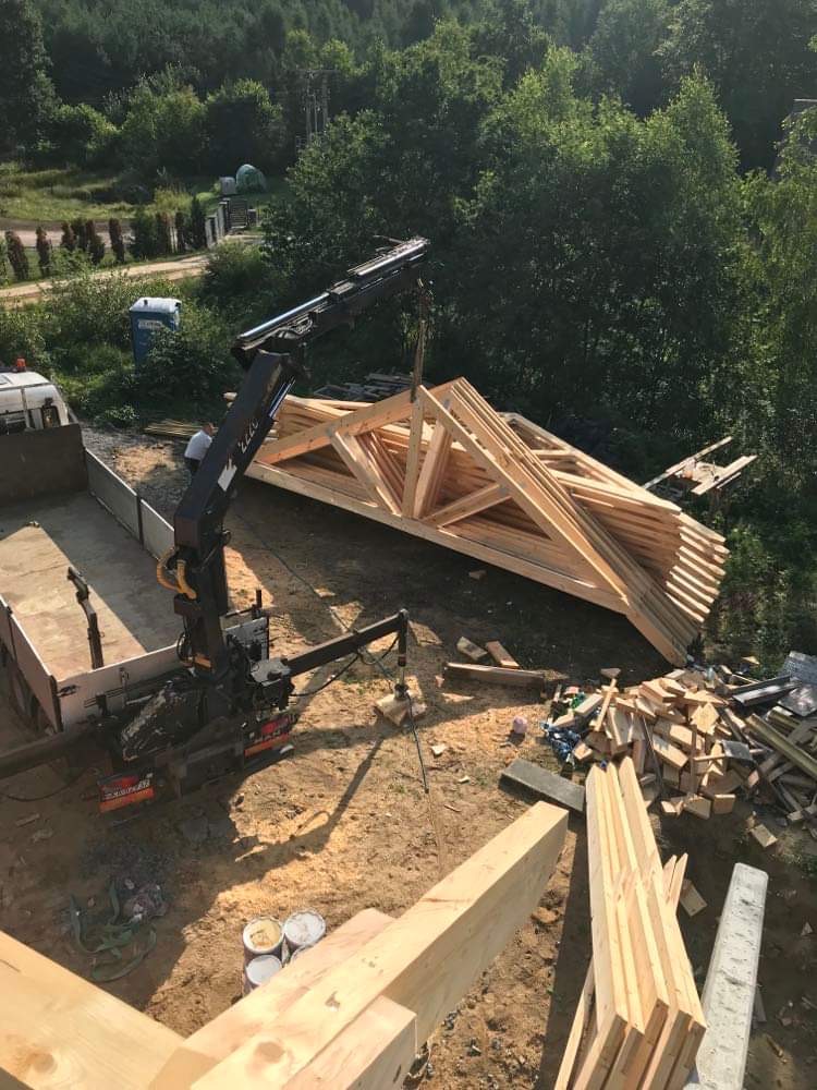 Montaż drewnianej konstrukcji dachu za pomocą dźwigu na placu budowy, widoczne elementy drewniane i narzędzia pracy.