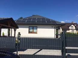 Dom jednorodzinny z panelami słonecznymi na dachu, widok zza metalowego ogrodzenia w słoneczny dzień.