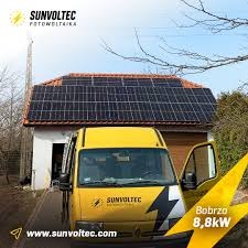 Instalacja paneli słonecznych na dachu budynku gospodarczego, widoczny żółty samochód dostawczy firmy Sunvoltec z logo i adresem strony internetowej, w tle słup energetyczny.