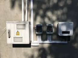 Szara ściana z zamontowaną białą skrzynką elektryczną z symbolem ostrzegawczym, dwoma licznikami i falownikiem. Białe rury biegną wzdłuż ściany. Na ścianie widoczne cienie drzew.