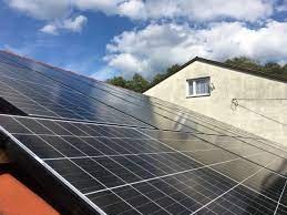 Panele słoneczne na dachu domu jednorodzinnego, widoczne odbicia chmur na powierzchni paneli, z boku fragment budynku z oknem.