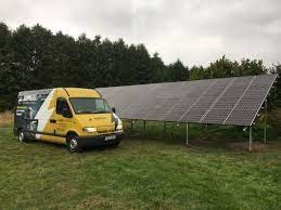 Żółty samochód dostawczy z grafiką firmy zaparkowany na trawie obok paneli słonecznych ustawionych na metalowej konstrukcji. W tle drzewa i pochmurne niebo.