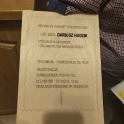 Tabliczka informacyjna z danymi prywatnego gabinetu ortopedycznego, wykonana na jasnym drewnie z ozdobnym obramowaniem w kształcie zygzaka, leżąca na stole obok paragonu i fragmentu koszuli w kratę.