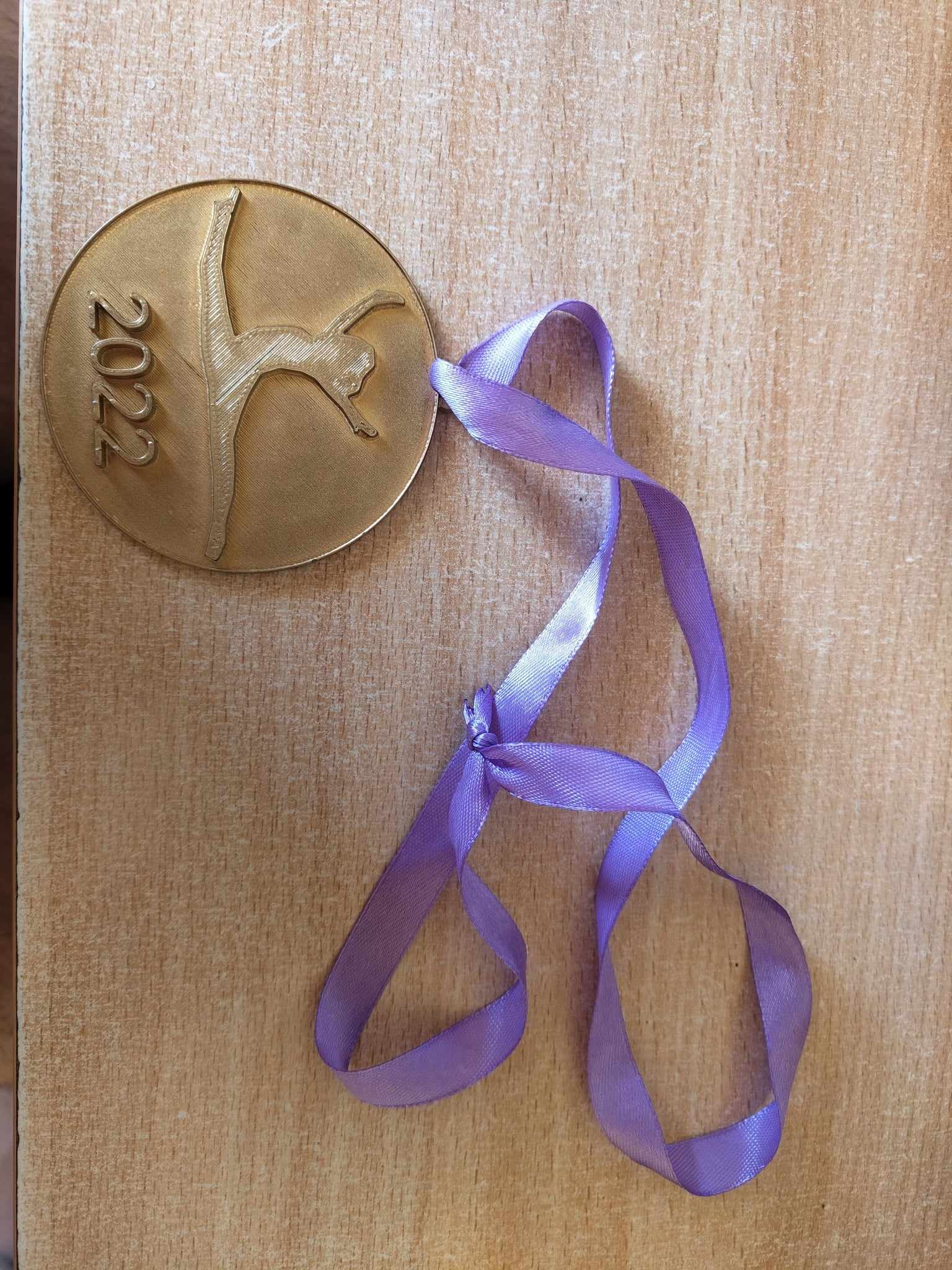 Złoty medal z wygrawerowaną sylwetką gimnastyczki i rokiem 2022, zawieszony na fioletowej wstążce, leżący na drewnianym blacie.