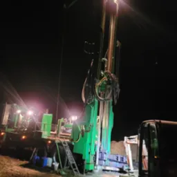 Wiertnica do studni głębinowych oświetlona reflektorami podczas nocnej pracy, widoczna drabina i fragment koparki.