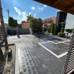 Nowoczesny podjazd z ciemnej kostki brukowej z białymi obramowaniami, widok zza drewnianej konstrukcji, w tle dom z minimalistyczną architekturą i starannie przycięte drzewa.