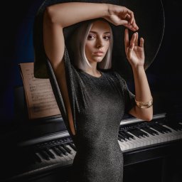 Archon studio fotograficzne Koszalin - Elegancki portret kobiety o platynowych włosach w szerokim kapeluszu i błyszczącej sukience, pozującej z uniesionymi dłońmi przy pianinie z nutami, w dramatycznym oświetleniu.