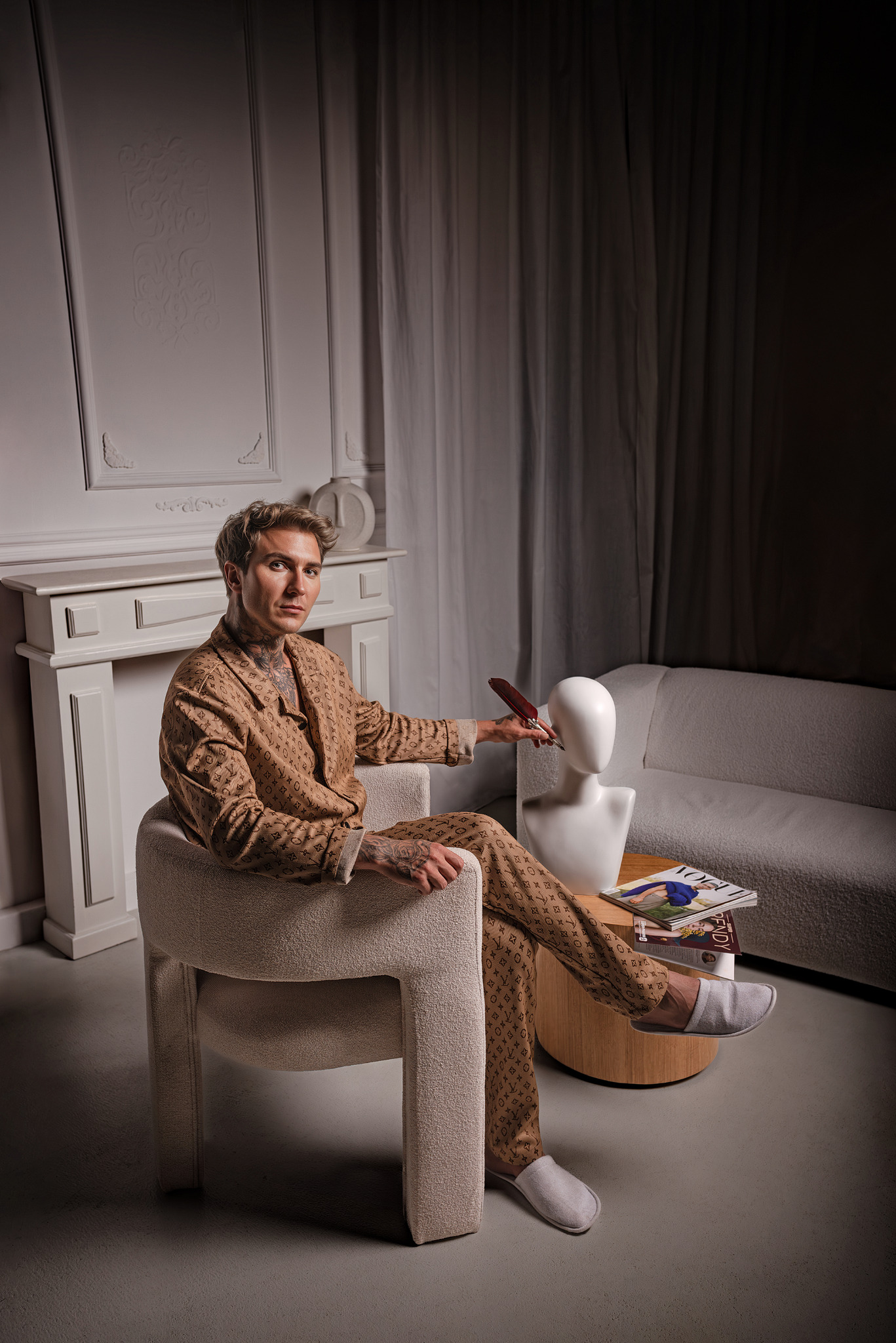 Mężczyzna w piżamie w monogramy siedzi w fotelu, trzymając pióro nad manekinem głowy, obok stolik z magazynami, w tle kominek i sofa.