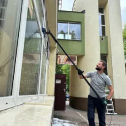 Mężczyzna z uśmiechem myje okna budynku za pomocą szczotki teleskopowej. Widoczne mydliny i woda spływająca po szybach. W tle budynek z elementami architektury.