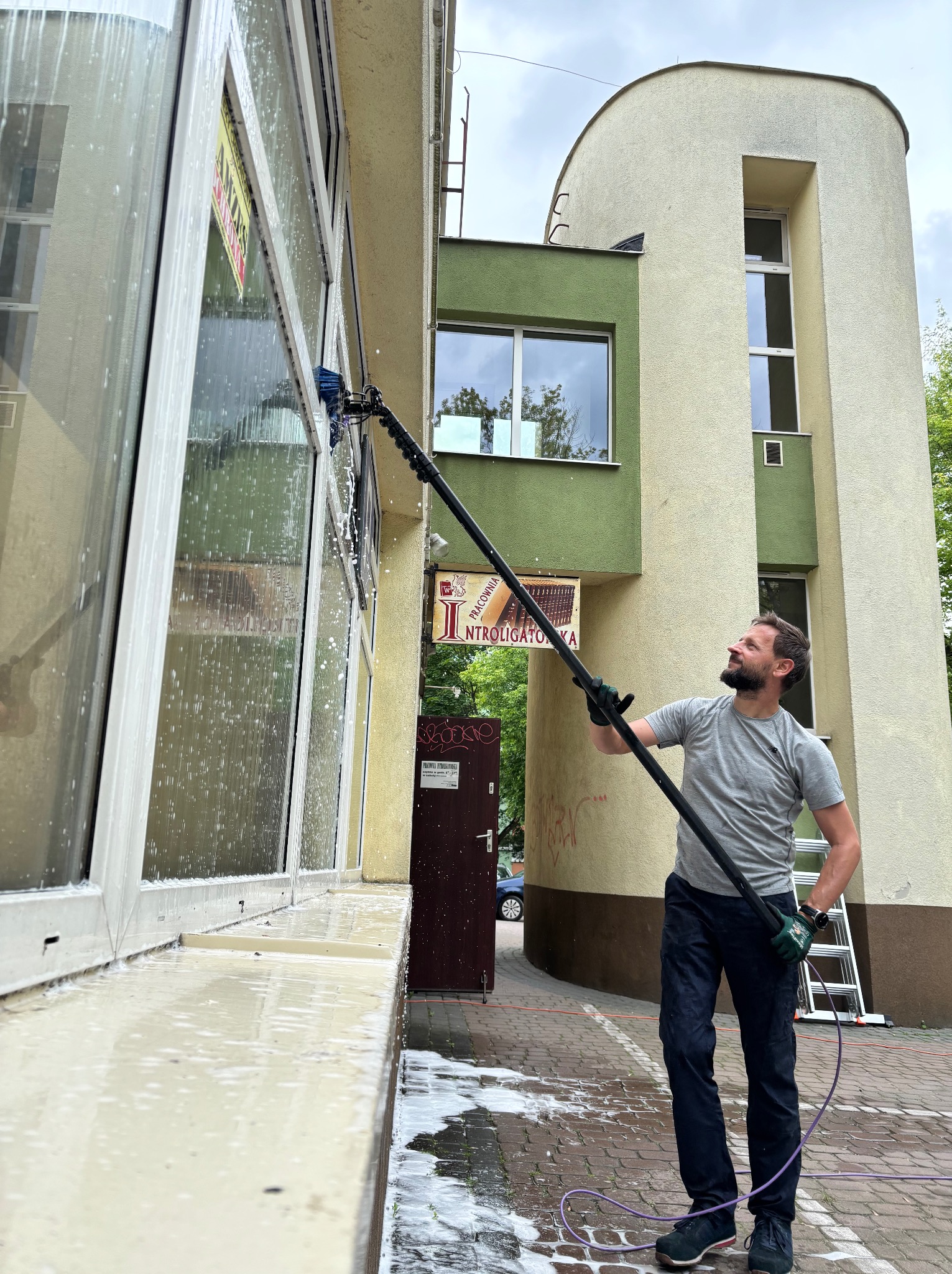 Mężczyzna z uśmiechem myje okna budynku za pomocą szczotki teleskopowej. Widoczne mydliny i woda spływająca po szybach. W tle budynek z elementami architektury.