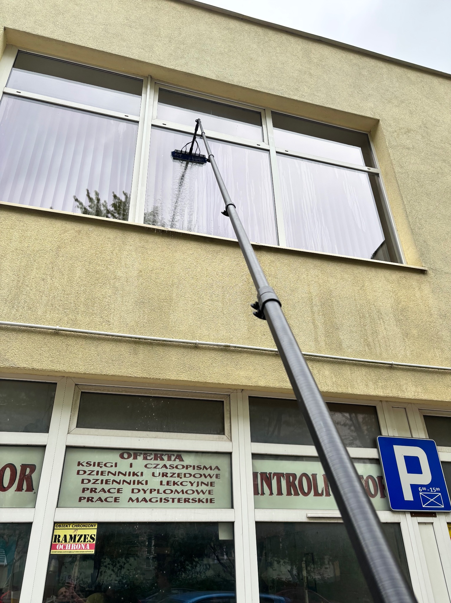 Zewnętrzne okno budynku myte teleskopową szczotką. Widoczne ślady wody spływającej po szybie. W tle elewacja i okna lokalu z ofertą usług.