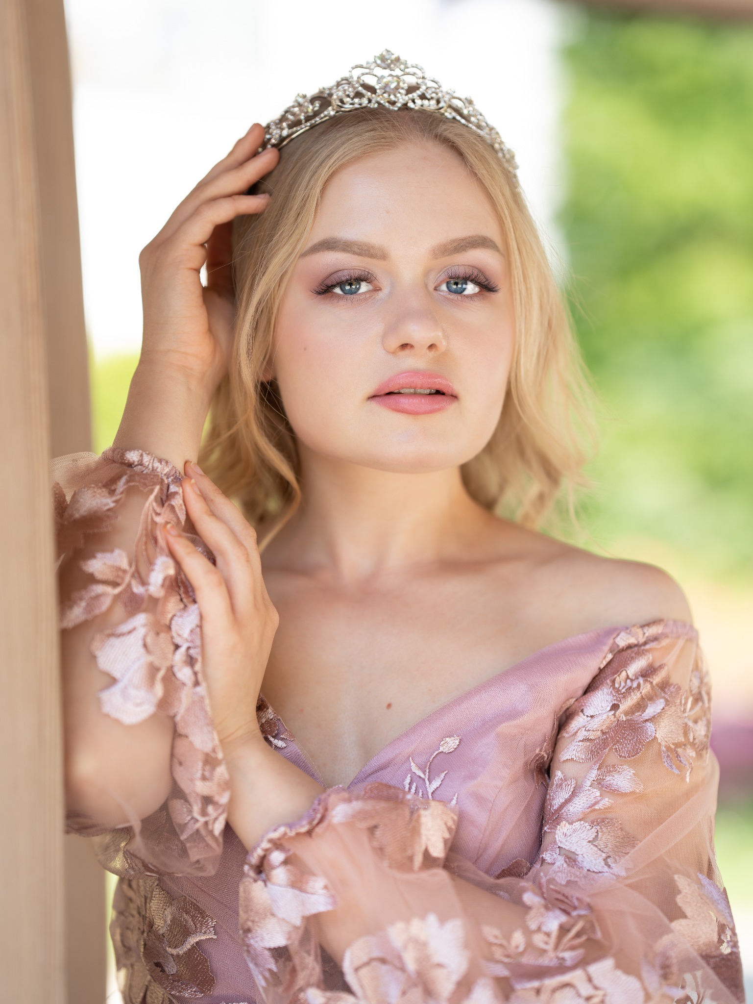 Portret młodej kobiety z tiarą, w zwiewnej sukni z kwiatowymi aplikacjami, pozującej na tle zieleni. Subtelny makijaż, naturalne światło.