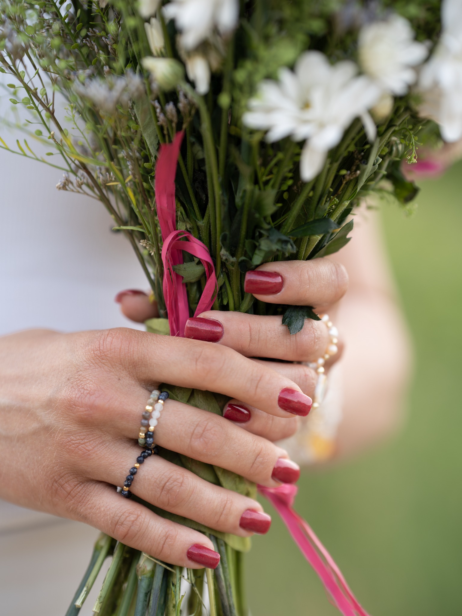 Dłonie kobiety z bordowym manicurem trzymają bukiet białych kwiatów, ozdobione pierścionkami z koralików i różową wstążką.