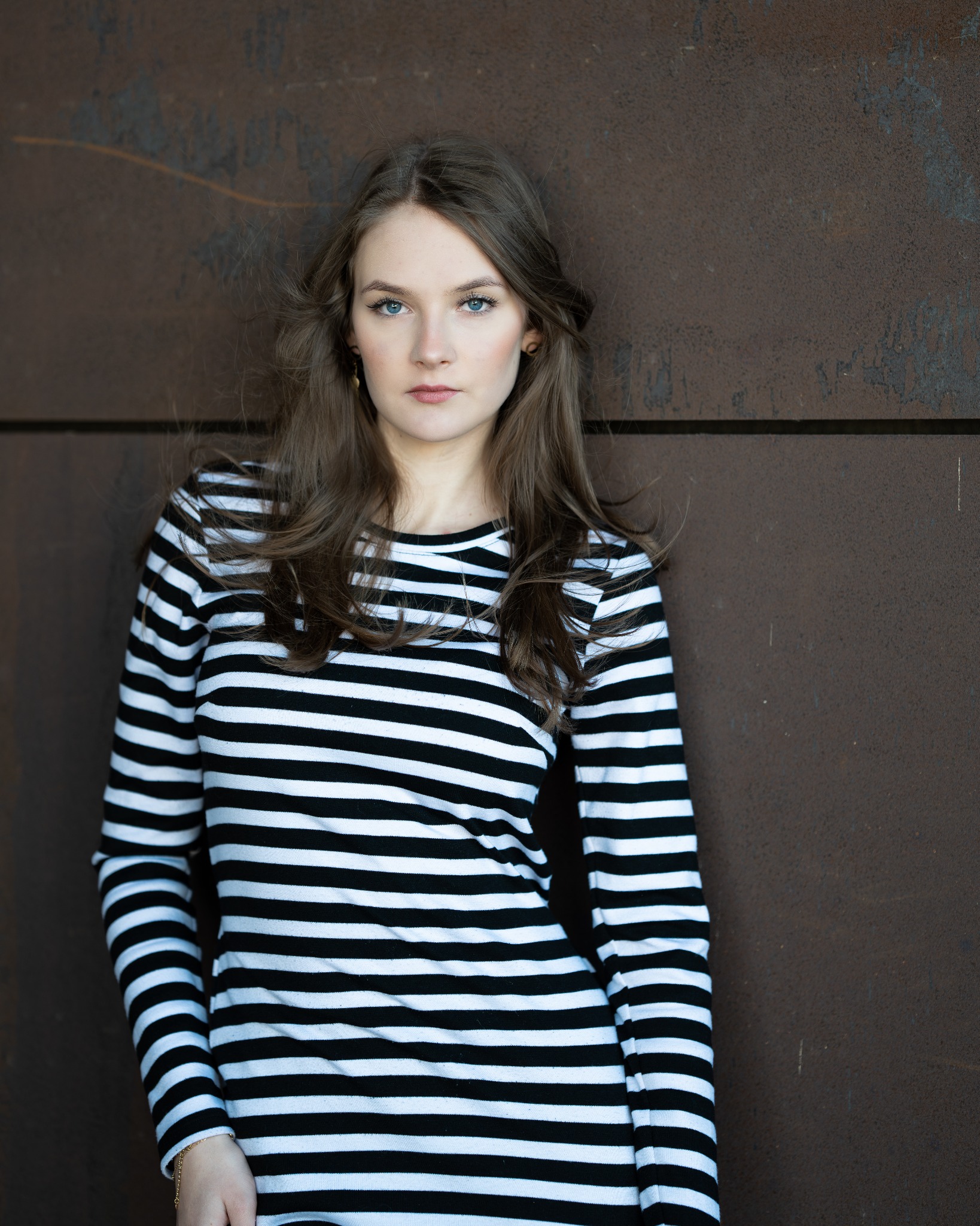 Portret kobiety w czarno-białej bluzce w paski, opierającej się o rdzawą ścianę, z wiatrem we włosach i subtelną biżuterią.