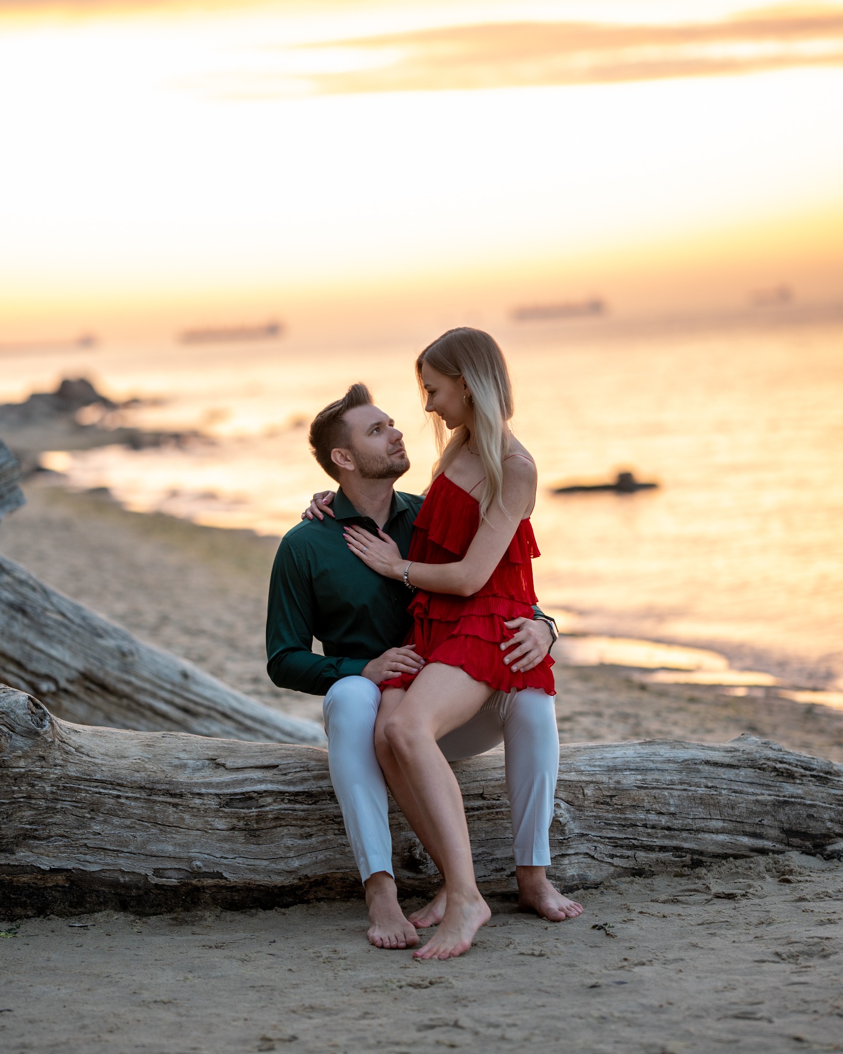 Zakochana para siedzi na pniu drzewa na plaży o zachodzie słońca, kobieta w czerwonej sukience spoczywa na kolanach mężczyzny w zielonej koszuli, oboje patrzą na siebie z czułością.