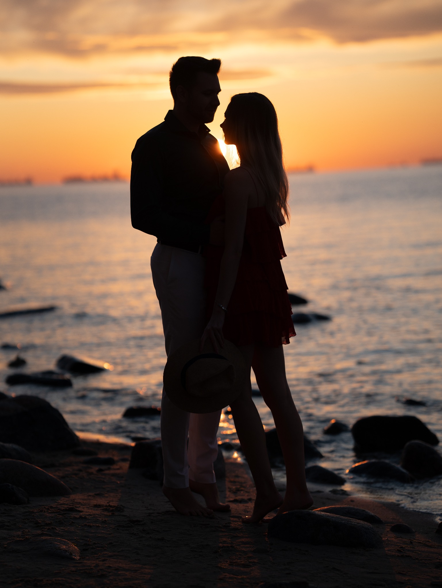 Sylwetki pary na plaży o zachodzie słońca, on w koszuli i spodniach, ona w sukience, trzyma kapelusz.