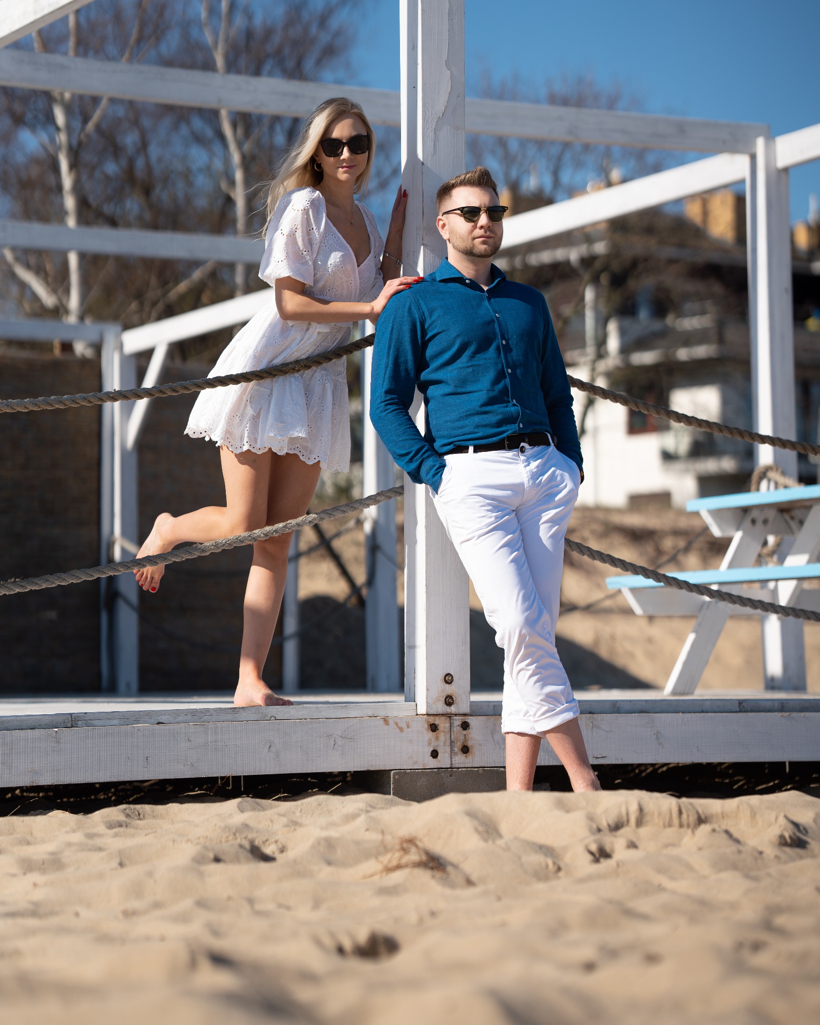 Elegancka para w okularach przeciwsłonecznych pozuje na plaży przy białej drewnianej konstrukcji. Kobieta w białej sukience, mężczyzna w niebieskiej koszuli i białych spodniach.