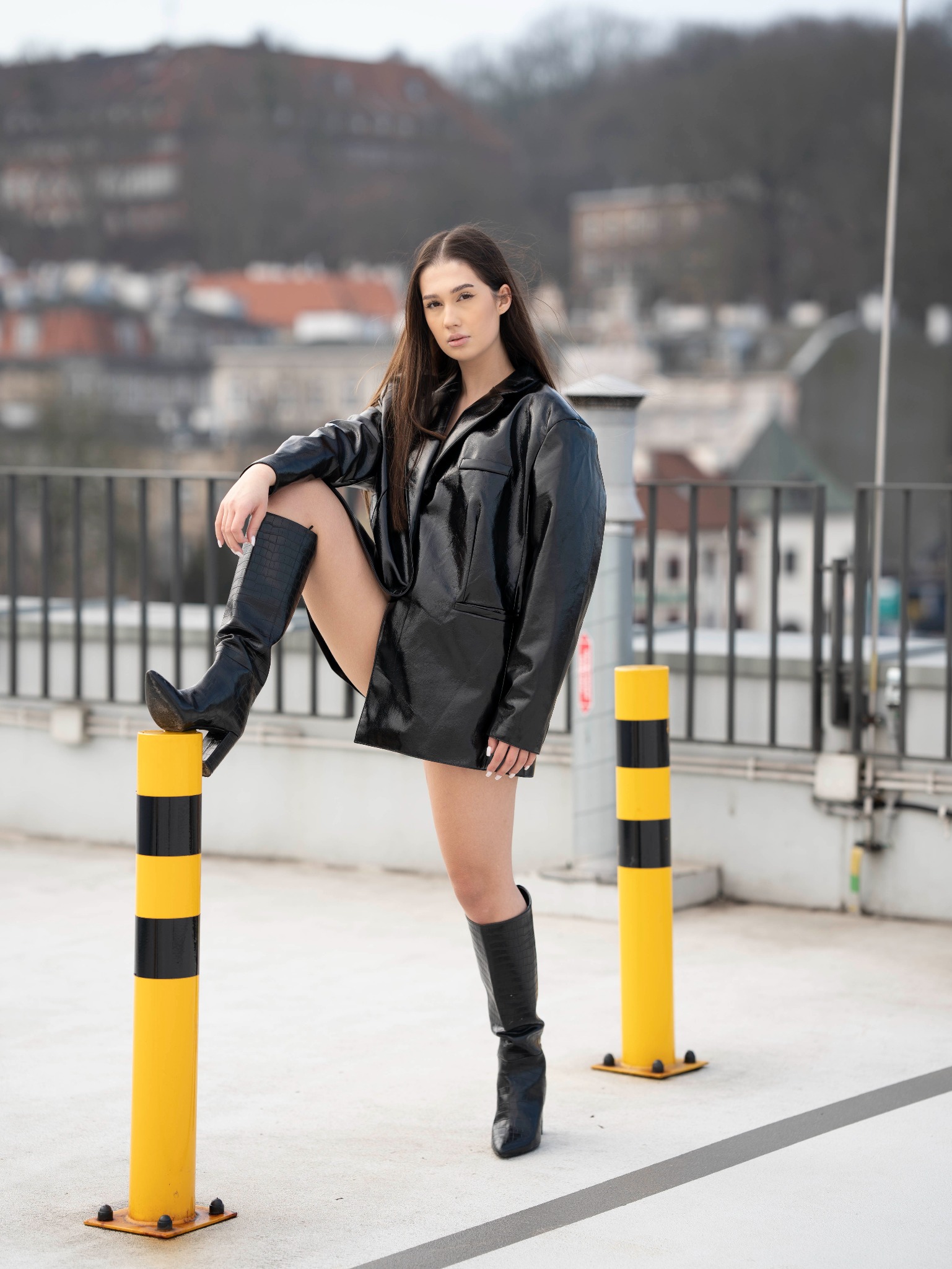 Kobieta w czarnej, lakierowanej kurtce i kozakach pozuje na dachu, opierając nogę o żółto-czarny słupek.