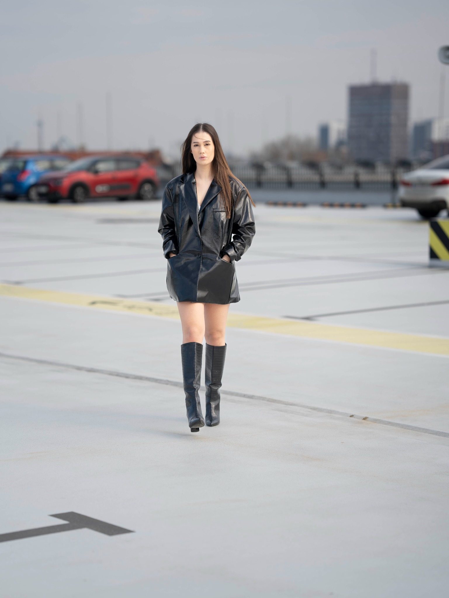 Kobieta w czarnej skórzanej sukience i kozakach pozuje na tle parkingu wielopoziomowego z widokiem na miasto w oddali.