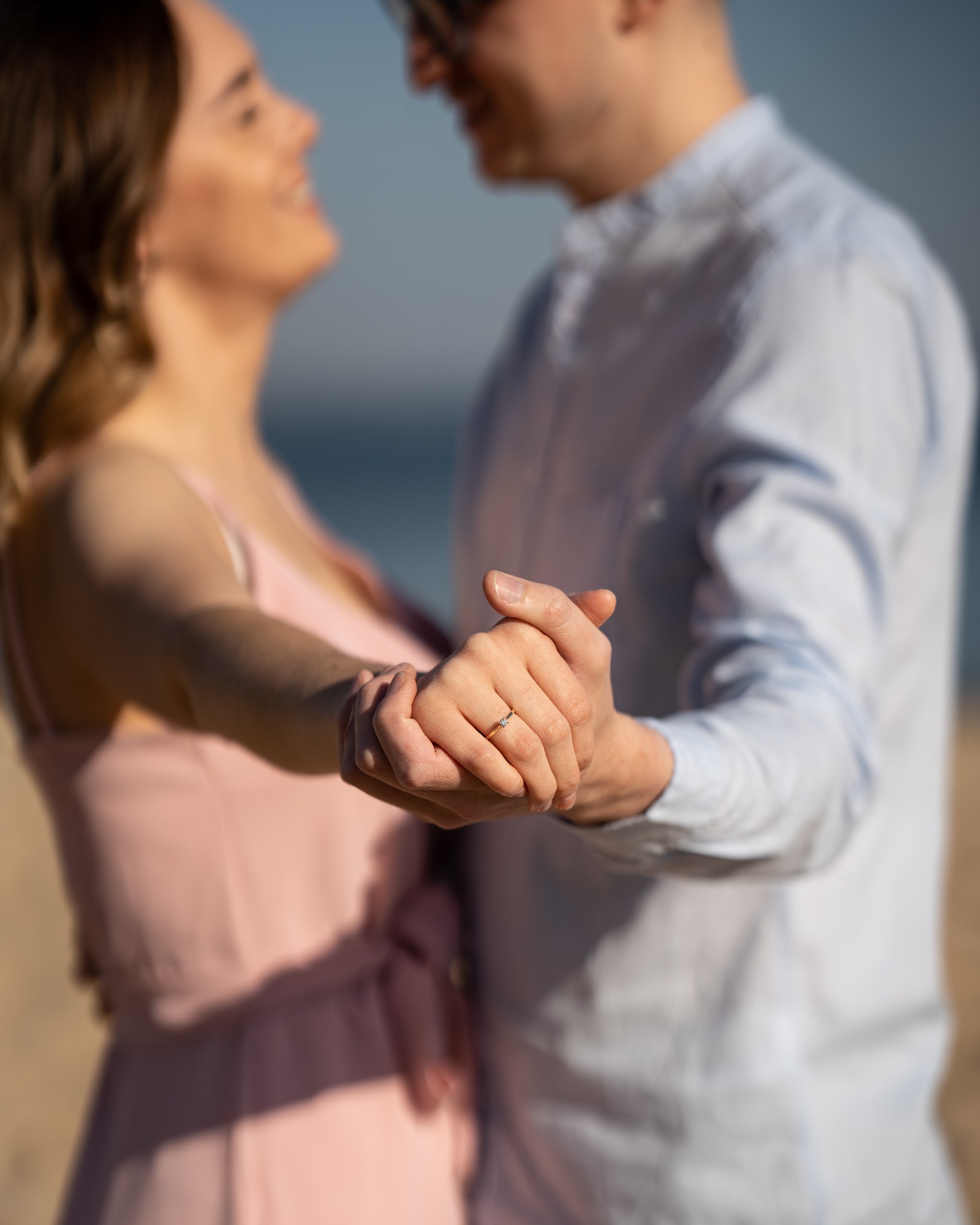 Para trzymająca się za ręce na tle plaży i morza, z widocznym pierścionkiem zaręczynowym na palcu kobiety.