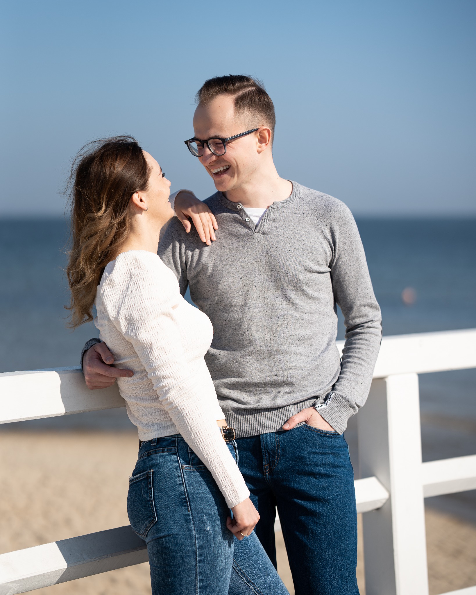 Zakochana para w Gdańsku, oparta o białą barierkę na tle morza, mężczyzna w okularach uśmiecha się do kobiety.