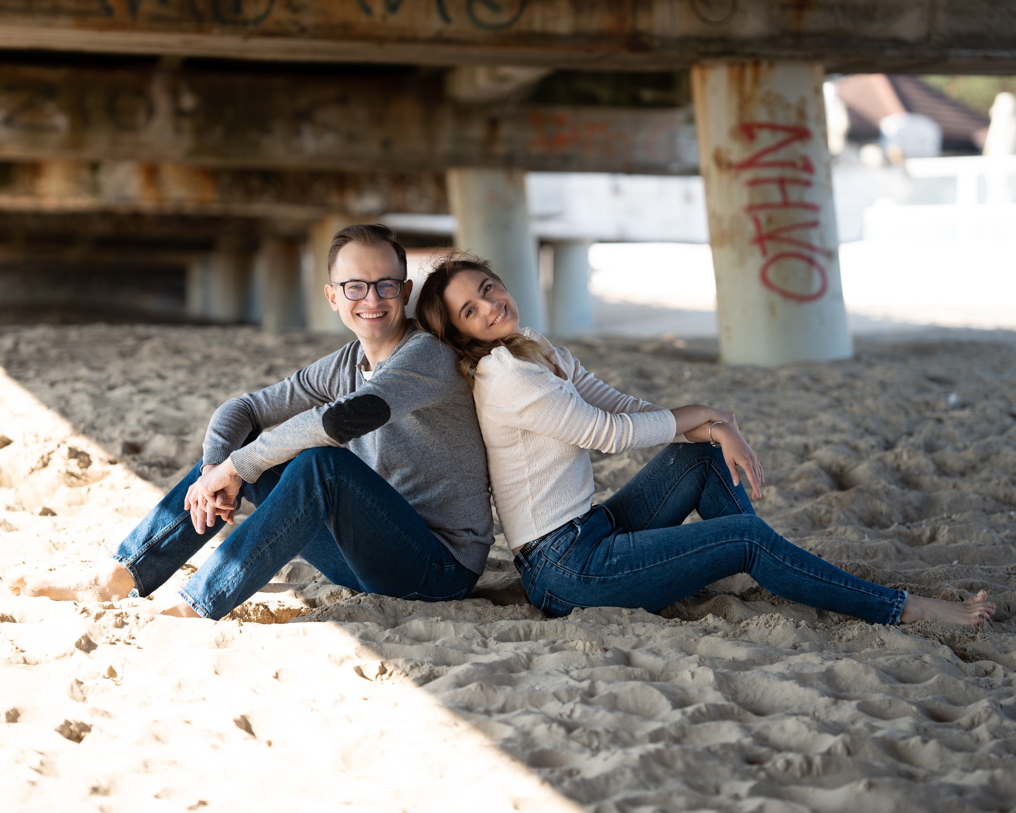 Uśmiechnięta para siedzi plecami do siebie na piaszczystej plaży pod betonowym pomostem z graffiti, w słoneczny dzień.