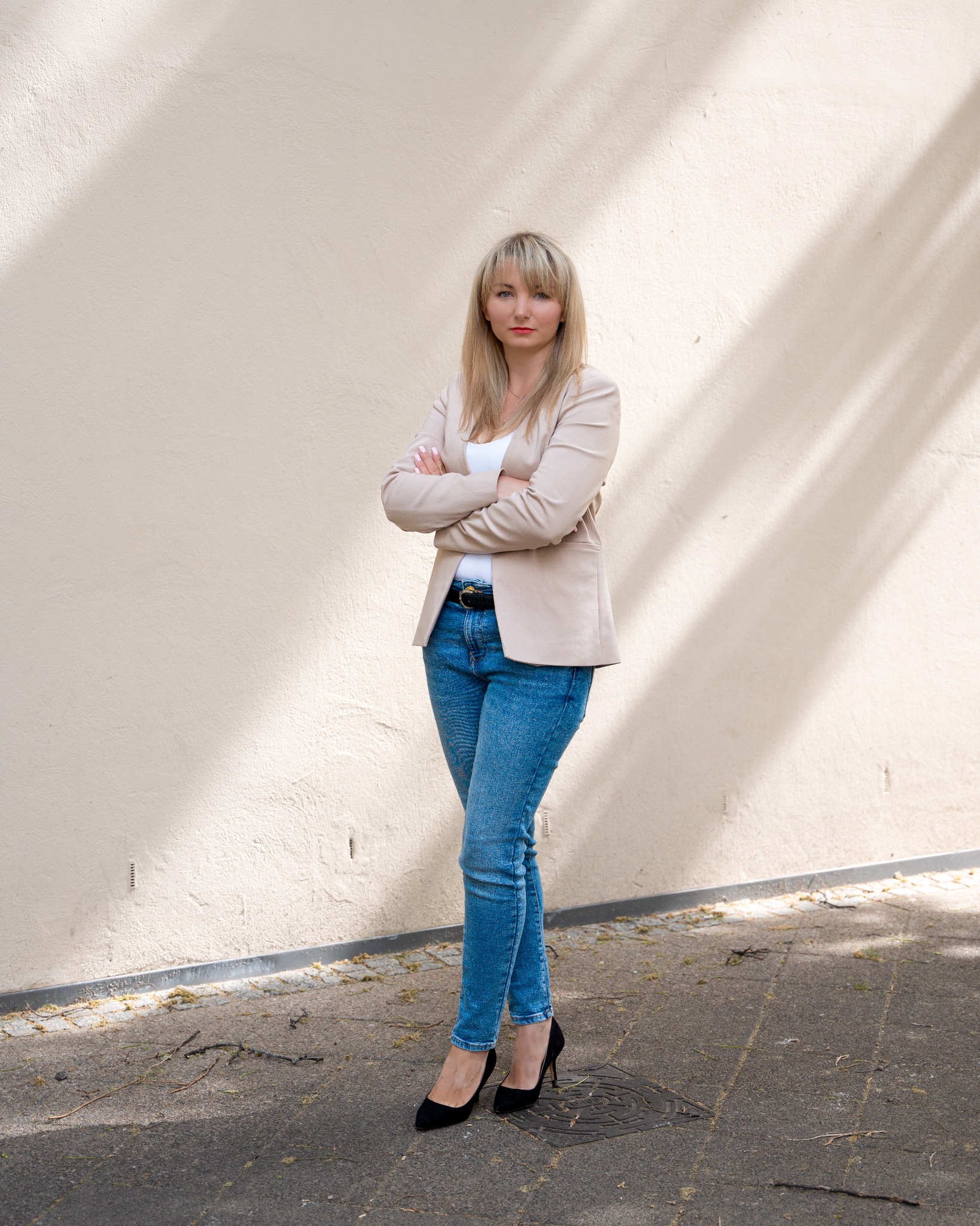 Portret kobiety w beżowej marynarce, jeansach i czarnych szpilkach, stojącej na tle kremowej ściany w słoneczny dzień.