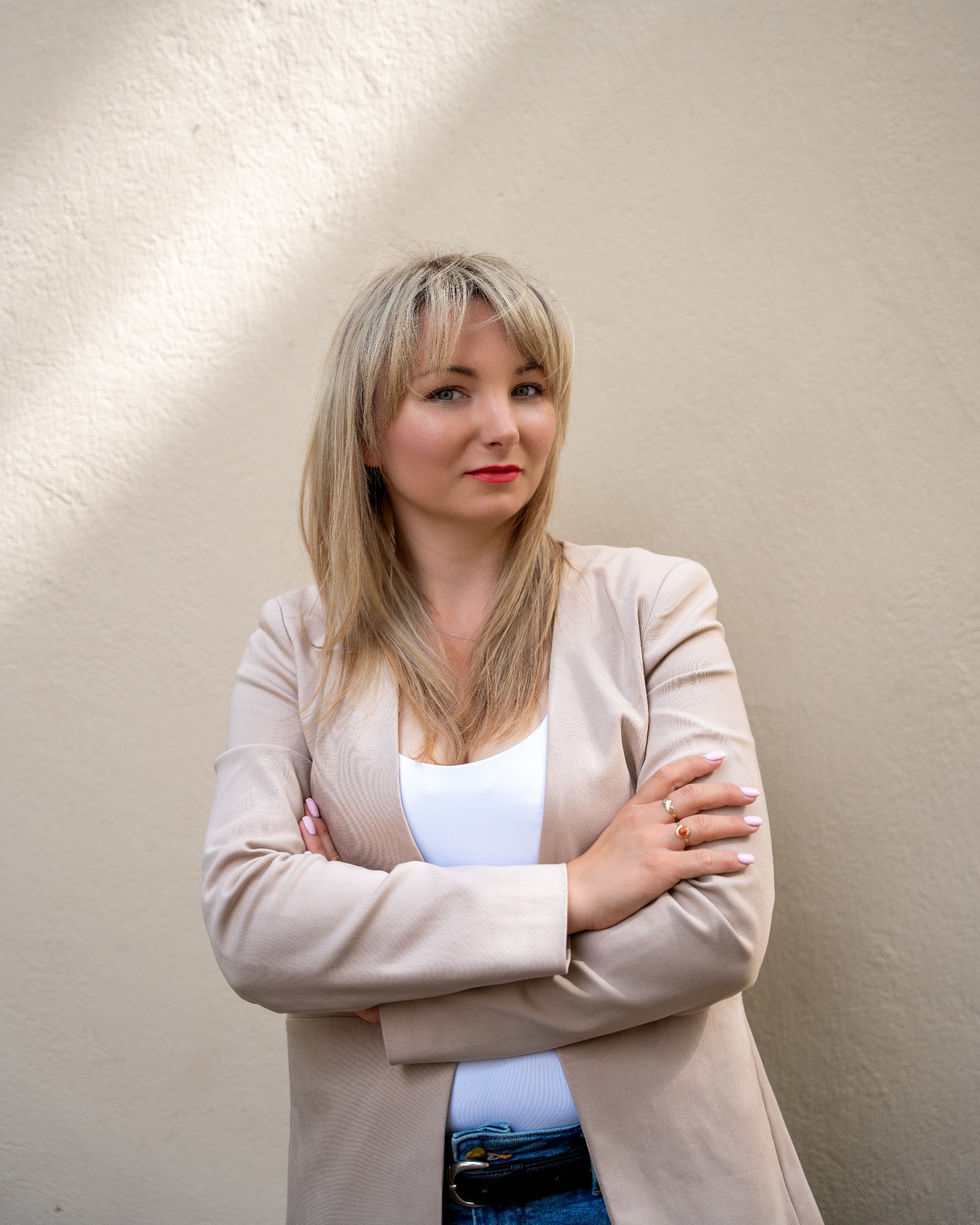 Portret kobiety z blond włosami, ubrana w beżową marynarkę, pozująca na tle jasnej ściany z delikatnym cieniem, skrzyżowane ręce, widoczne pierścionki na palcach.