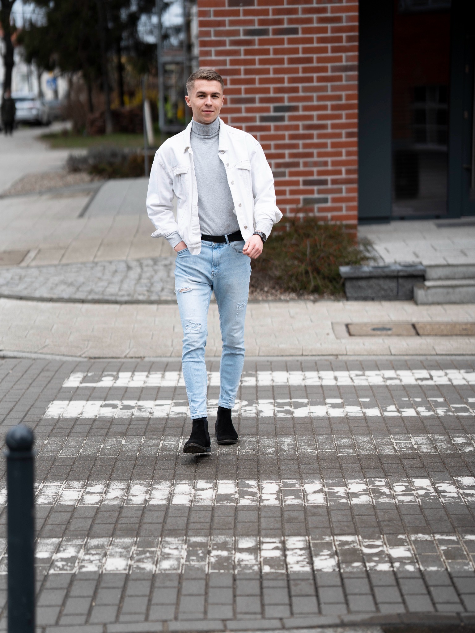 Mężczyzna w jasnej kurtce jeansowej i podkoszulku z golfem przechodzi przez pasy na ulicy, z rękami w kieszeniach, na tle budynku z czerwonej cegły.