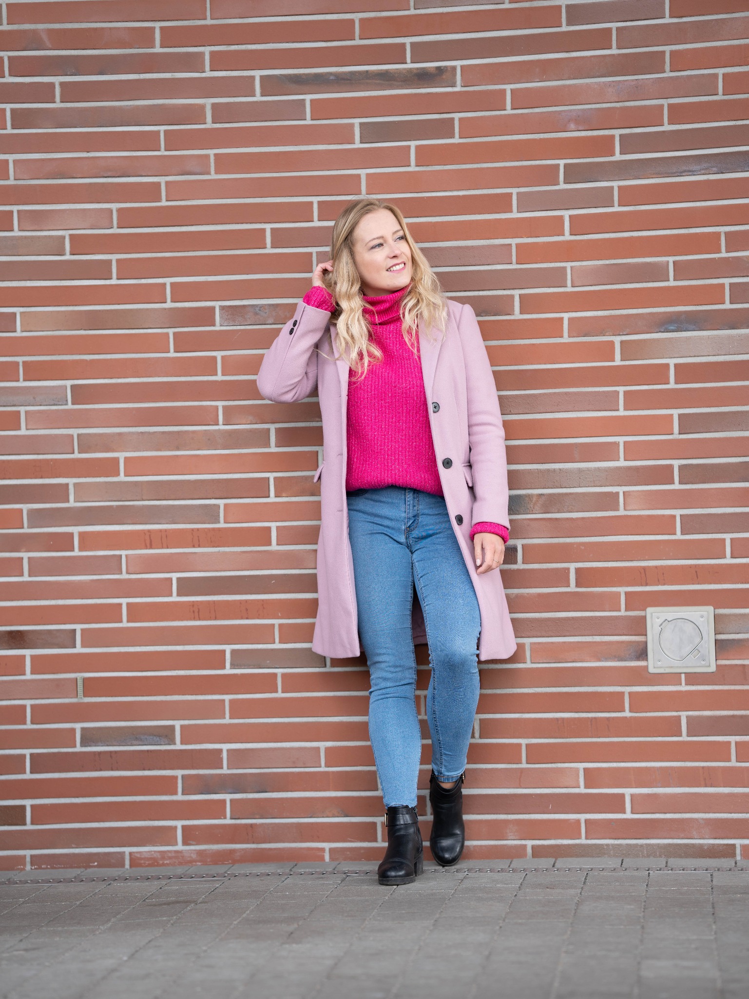 Portret kobiety w różowym płaszczu i swetrze, opierającej się o ceglaną ścianę, z delikatnym uśmiechem i dłonią we włosach.