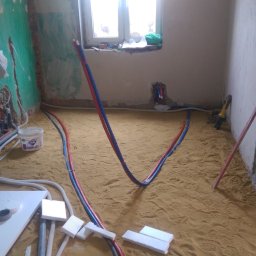 PROBUDOTECH - Wnętrze w trakcie remontu: podłoga pokryta piaskiem, wystające rury wodne (czerwone i niebieskie), przewody elektryczne i narzędzia w tle.