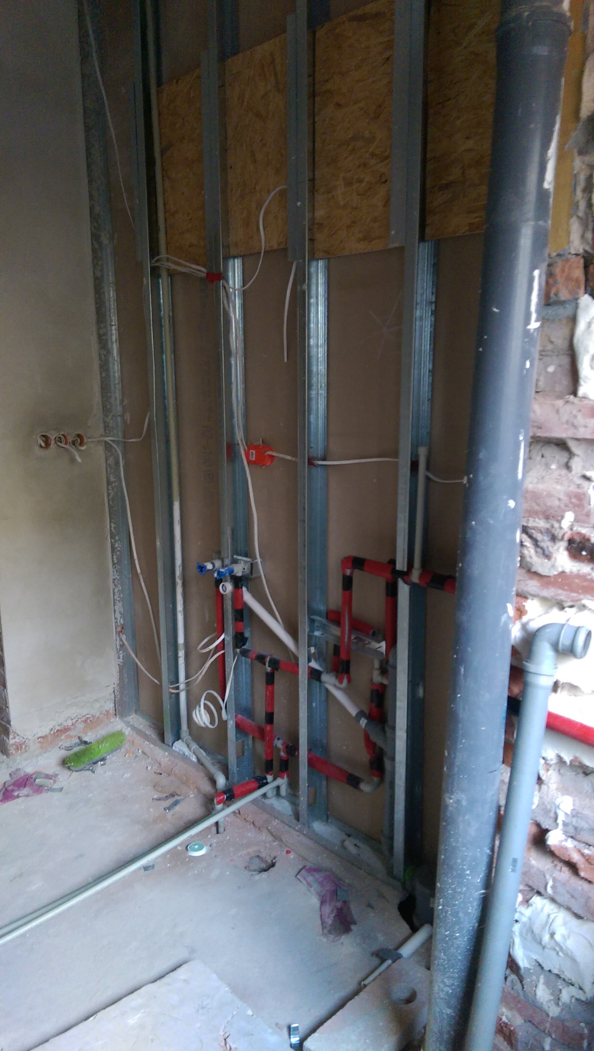 Instalacja wodno-kanalizacyjna w trakcie remontu łazienki: widoczne metalowe profile, rury z tworzywa w kolorze czerwonym i czarnym, wystające przewody elektryczne i fragment ściany z cegły