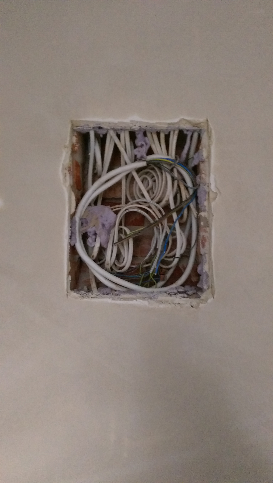 Otwór w ścianie z widocznymi przewodami elektrycznymi zabezpieczonymi pianką montażową, cegła w tle.
