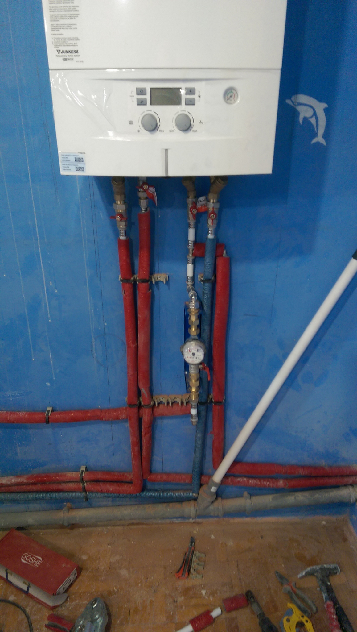 Biały piec gazowy marki Junkers zamontowany na niebieskiej ścianie z rurami w izolacji, widoczne narzędzia i elementy instalacji na podłodze.