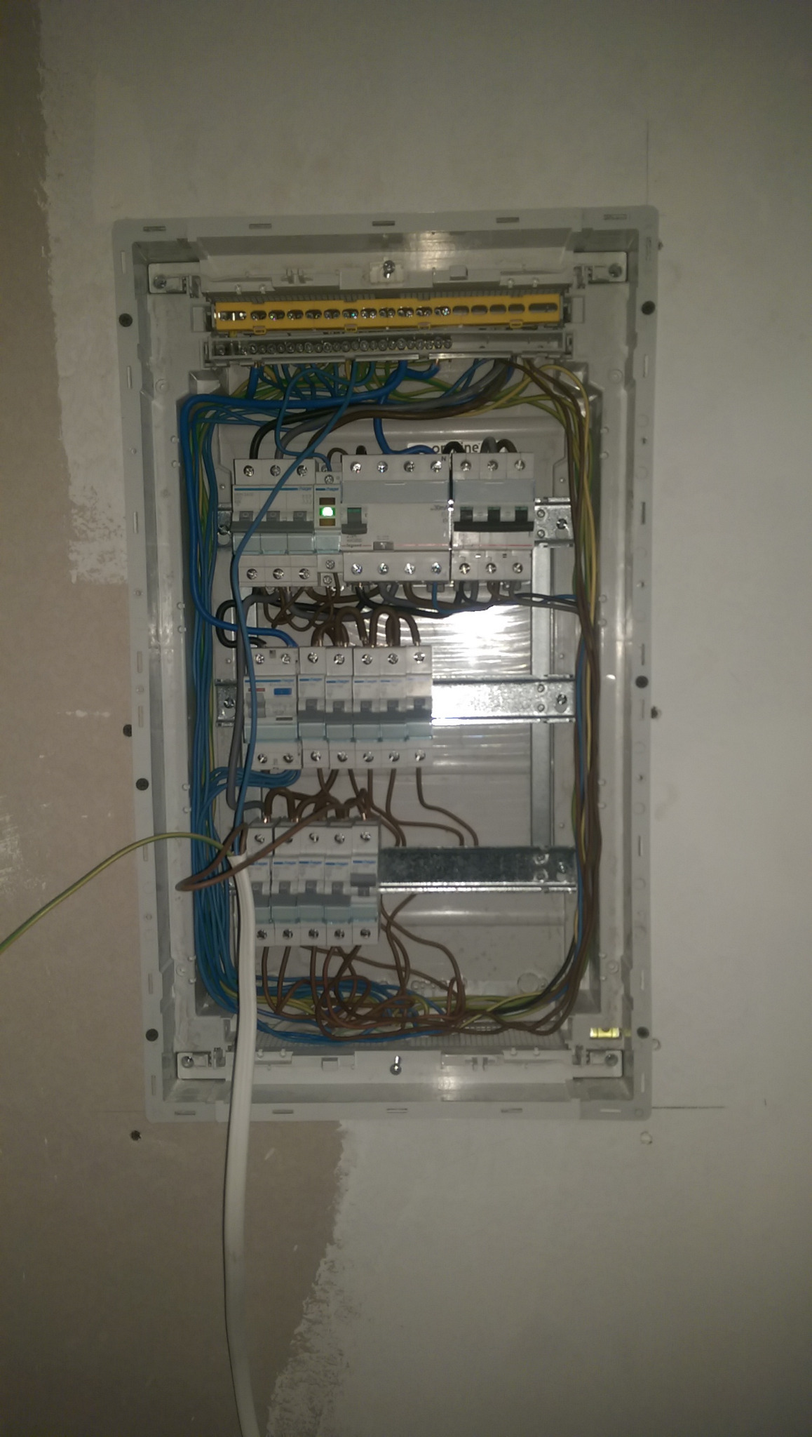 Rozdzielnia elektryczna z bezpiecznikami, widoczne kable w kolorach niebieskim, brązowym i żółto-zielonym, zamontowana w ścianie przed malowaniem.