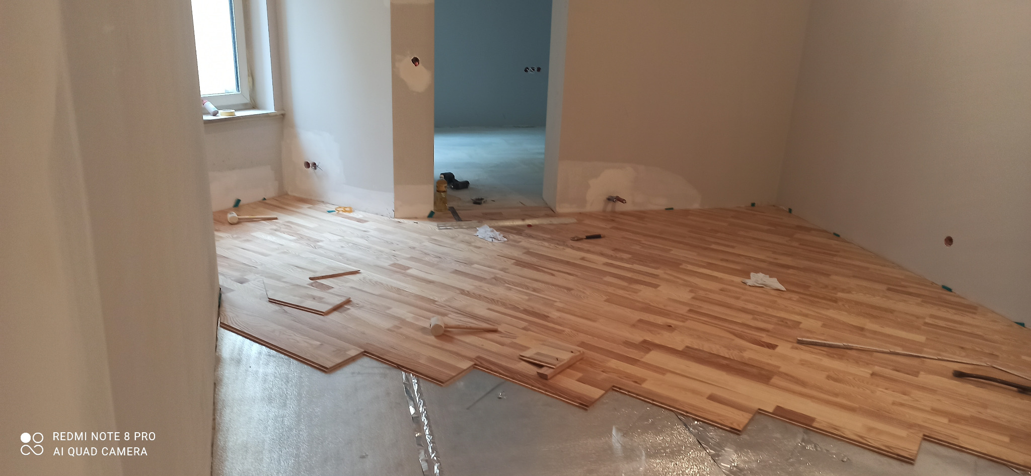 Układanie parkietu z jasnego drewna w nowym mieszkaniu, widoczne fragmenty folii izolacyjnej, młotek gumowy i niedokończone fragmenty podłogi.