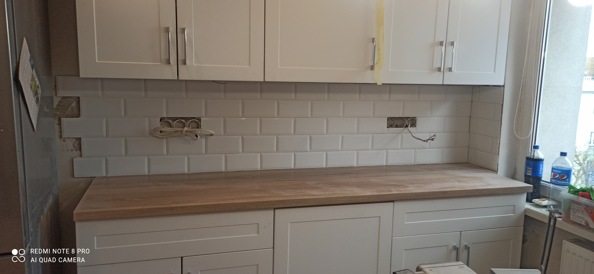 Fragment kuchni w trakcie remontu: białe szafki wiszące i stojące, drewniany blat, białe płytki na ścianie z widocznymi puszkami elektrycznymi i wystającymi przewodami, częściowo położone płytki...