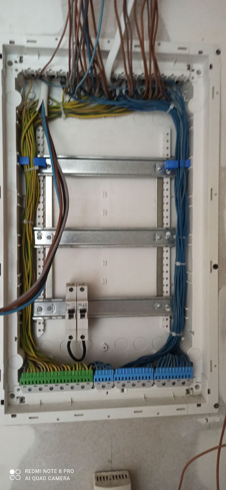 Rozdzielnica elektryczna z zamontowanymi przewodami w kolorach: brązowym, niebieskim, żółto-zielonym, zabezpieczonymi niebieskimi opaskami zaciskowymi i podłączonymi do listew zaciskowych...