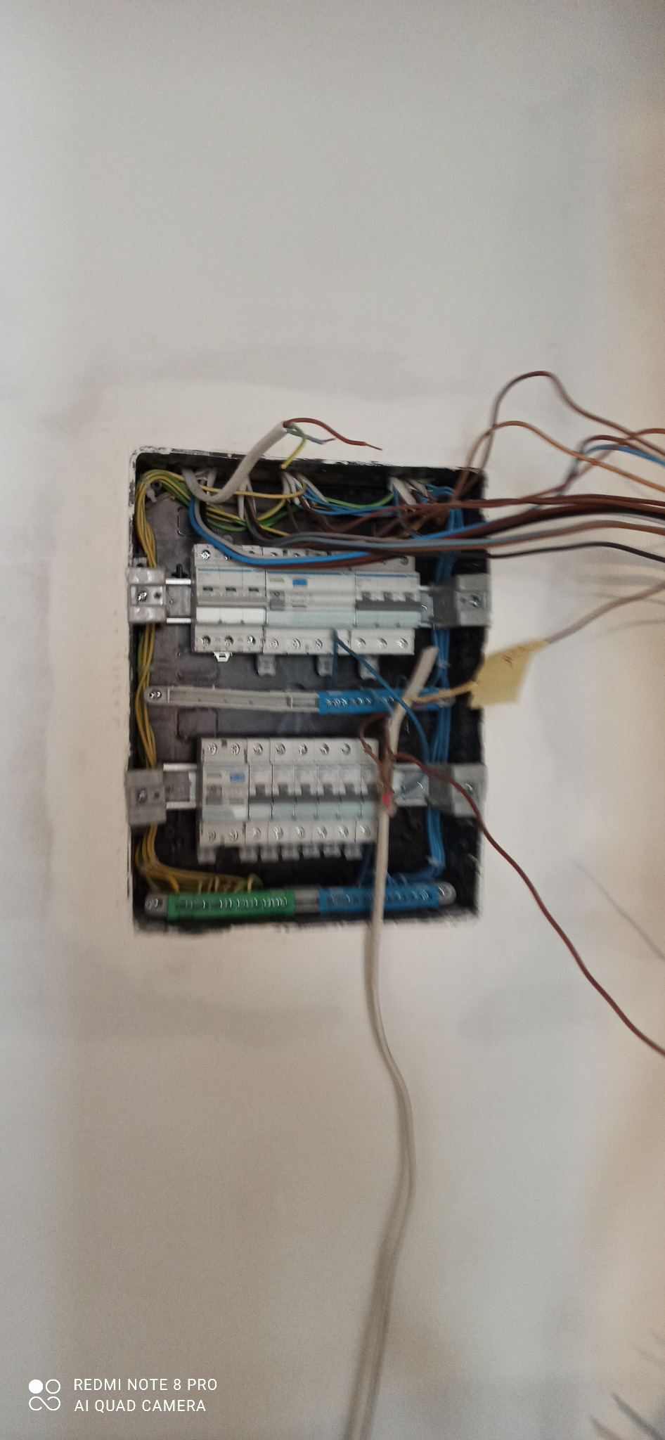 Otwarta skrzynka elektryczna z widocznymi bezpiecznikami i kablami o różnych kolorach na tle białej ściany.