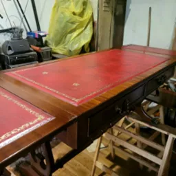 Odrestaurowane biurko z czerwonym blatem obitym skórą i złotym zdobieniem, widoczne szuflady z ozdobnymi uchwytami, w tle warsztat z narzędziami i folią ochronną.