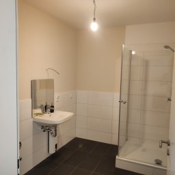 Olbut - Nowoczesna łazienka z białymi płytkami, ciemną podłogą, kabiną prysznicową ze szklanymi drzwiami i umywalką z lustrem, oświetlona pojedynczą żarówką wiszącą na kablu.