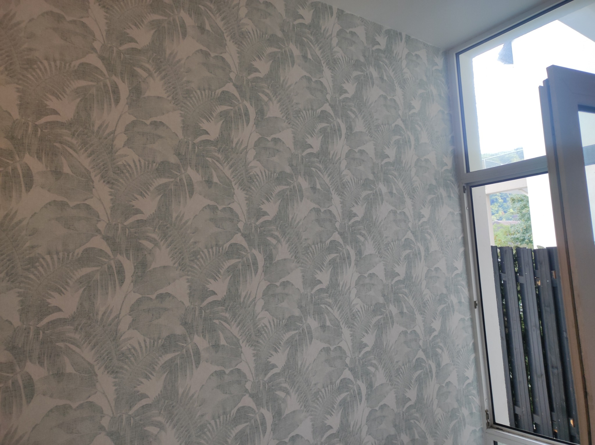 Ściana pokryta tapetą w jasne, stonowane wzory liści palmowych, widoczna z bliska, z fragmentem białej ramy okiennej po prawej stronie, przez którą widać fragment szarego ogrodzenia i zieleni.